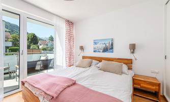 A bright bedroom with two single beds and a modern wooden headboard. Large windows provide plenty of daylight and a view of a balcony.