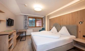 A modern hotel room with a large double bed and wooden paneling. The room features a window, a desk, and a television.