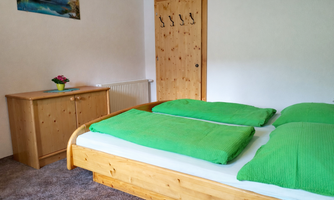 A simple bedroom with a wooden bed and green cushions. A picture hangs on the wall, and there is a wooden table with a plant.