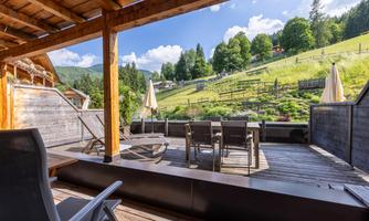 A cozy terrace with garden furniture and sun umbrellas. In the background, green hills and trees are visible.