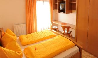A cozy bedroom with two single beds and orange bedcovers. There is a desk and a television on the wall.