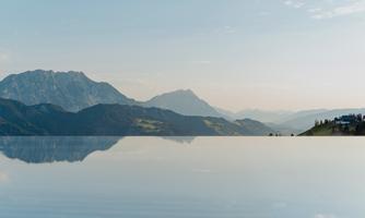 A tranquil mountain landscape with gentle hills and majestic mountains in the background. The sky is clear and is reflected in a calm water surface.