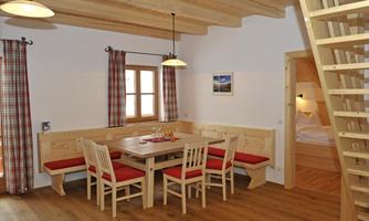 A cozy dining area with a wooden table and chairs in a bright room. The decor features rustic details and a staircase leading to an upper area.