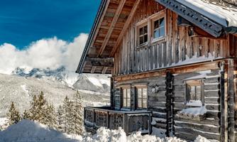 A cozy wooden house in the snow with a view of the mountains. The sky is clear and the landscape is wintery and snowy.