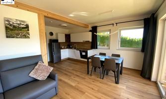A modern kitchen with a dining area and a cozy living area. Large windows provide plenty of daylight and a view of the greenery.