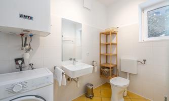 A bright bathroom with a washing machine and a hand wash basin. On the right side, there is a toilet and an open shelf.