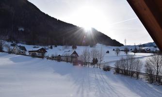 A snowy landscape with hills and a radiant sunrise. In the foreground, there are a few houses and bare trees.