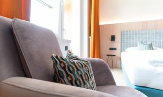 A cozy armchair with a decorative cushion. In the background, a bright bedroom with a bed and large windows is visible.