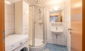 A modern bathroom with a shower, a sink, and a washing machine. The walls are tiled, and there is a large mirror.