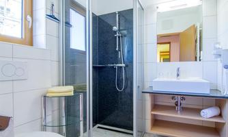 A modern bathroom with a shower, a sink, and a window. The walls are brightly tiled, and there are open shelves for towels.