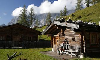 A picturesque mountain cabin with green meadows and a clear sky. A person is sitting relaxed on a bench in front of the cabin.