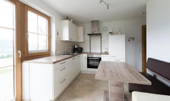 A modern kitchen with white cabinets and a wooden table.  
Plenty of light from large windows creates a friendly atmosphere.