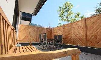 A cozy terrace with a wooden bench and a table with chairs. Surrounded by a wooden privacy screen and a clear blue sky.