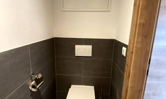 A small, modern restroom with gray tiles and a white toilet seat. The walls are partially tiled in light and partially in dark, and there is a toilet paper holder.