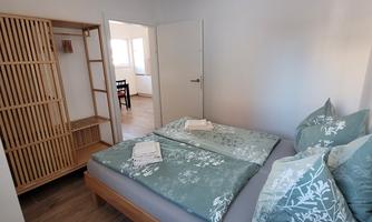 A cozy bedroom with two beds and decorative pillows. The room is bright and friendly, with a wooden wardrobe and access to another room.
