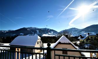 A winter landscape with snow-covered huts and mountains in the background. The sky is clear and adorned with flying hot air balloons.