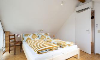 A cozy bedroom with a double bed and a chair. The walls are bright and there is plenty of natural light.