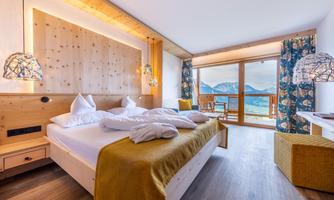 A modern room with a large bed and wooden details. Through the windows, one looks out at a picturesque mountain landscape.