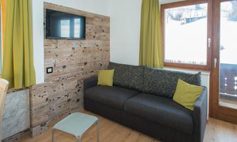 A cozy living room with a gray sofa and yellow cushions. There is a television on the wall and large windows with a view of the snow-covered landscape.