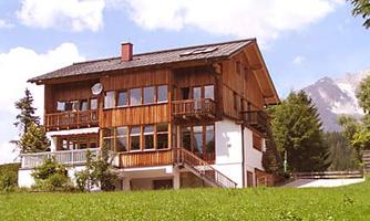 A charming, modern wooden house with large windows. In the background, trees and mountains are visible.