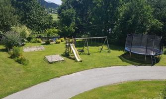 A beautiful garden with a slide, a swing, and a trampoline. Surrounded by trees and well-maintained grass.