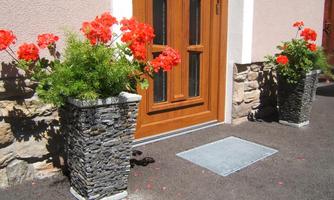 A beautiful entrance with a wooden door and colorful, red flowers in stone planters. The ground is paved with asphalt and the atmosphere is inviting.