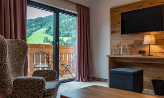 A cozy room with a large window offering a view of the green hills. There is a comfortable armchair, a table, and a flat-screen TV.