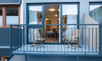 A small balcony with two wooden chairs and a view of a bright room. The windows are open and the curtains are gently flowing.