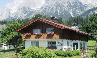 A charming alpine house with blooming windows and a well-kept garden. Majestic mountains rise in the background under a clear sky.