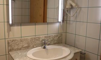 A simple bathroom with a sink on a marble surface. The mirror is equipped with integrated lighting, and the walls are tiled.