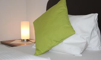 A cozy bed with a green pillow and white covers. Next to the bed is a lamp that gently glows.