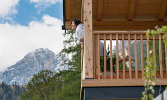 A cozy wooden house with a balcony, where a person stands enjoying the view. In the background, green forests and majestic mountains can be seen.