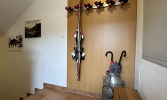 An entrance area with a wooden staircase, a wall with skis and decoration. On the right stands a milk canister and an umbrella.