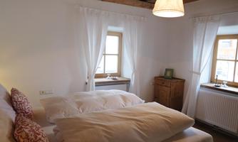 A cozy bedroom with a double bed and white bedspreads. The windows are adorned with light curtains and a wooden ceiling gives the room a rustic charm.