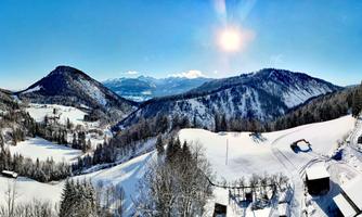 A winter landscape with snow-covered mountains and a bright blue sky. The sun shines brightly over the idyllic scene.