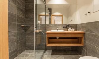 A modern bathroom with gray tiles and wooden details. It features a shower, a sink, and an elegant wooden cabinet.