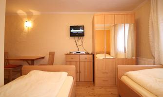 A cozy room with two single beds and a television on the wall. The room is bright and has a mirror and a wooden wardrobe.