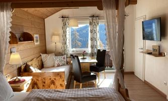 A cozy room with wooden decor and a double bed. There are seating areas, a table, and a beautiful view of the mountains.