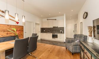 A bright and modern kitchen with a dining area and a cozy armchair. The room has a warm atmosphere with wood and stylish decorative elements.