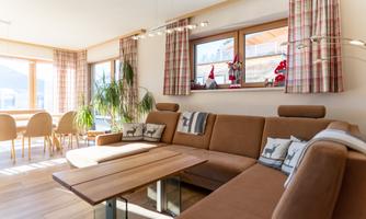 A cozy living room with a brown sofa and a wooden table. Large windows let in a lot of light and there are decorative plants and Christmas ornaments.