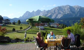A group of three people is sitting at a table outdoors, enjoying the view. In the background, majestic mountains and a green sun umbrella can be seen.