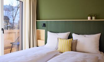 A modern bedroom with two single beds and green walls. Large windows let in plenty of light and offer a view outside.