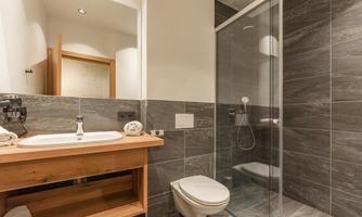 A modern bathroom with gray tiles and a glass shower cubicle. Towels and toiletries are placed on the wooden surface.