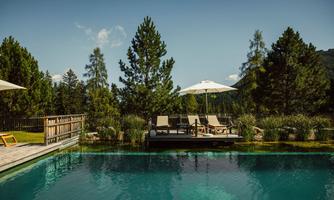 A picturesque pool surrounded by lush greenery and tall trees. Loungers with umbrellas invite relaxation.