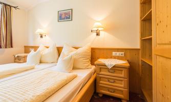 A cozy room with two single beds, wooden shelves, and warm light. The walls are light, and there is a small picture frame on the wall.