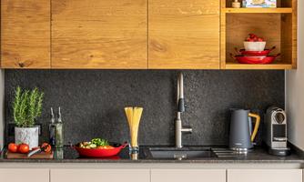 A modern kitchen with wooden shelves and a black countertop. Fresh vegetables, a kettle, and dishes are placed on the counter.