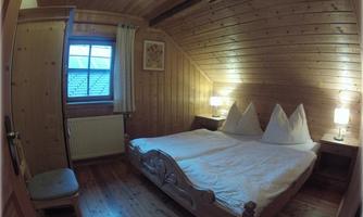 A cozy bedroom with wooden ceiling and walls. There is a double bed with white pillows and nightstands, as well as a chair in the corner.