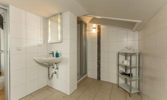 A modern bathroom with white tiles and a sink. In the background, a shower niche and a shelf can be seen.