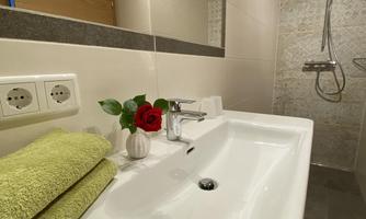 A modern bathroom with a white sink and a large mirror. On the shelf are green towels and a small vase with a red rose.