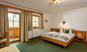 A cozy bedroom with a double bed, wooden furniture, and carpet flooring. Windows with curtains offer a view outside.